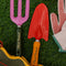 two metal gardening tools: red and yellow 'plant parent' towel and pink and green 'plant parent' trident on grass