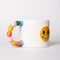 side view of white ceramic mug with rainbow shapes handle and yellow smiley face on the front
