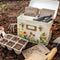cream metal seed box with floral and butterfly print on the bottom and three interior compartments in a garden with gardening tools