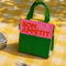 bubblegum, kelly green and yellow colorblock lunch bag with red bon appetit across the front on a yellow gingham blanket
