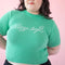 model wearing pale green short sleeve mockneck sweater with white cursive 'stay soft' across the front