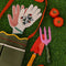 two metal gardening tools: red and yellow 'plant parent' towel and pink and green 'plant parent' trident on grass surrounded by gardening items