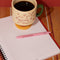 cream short ceramic mug with pink cursive 'in my growing season' with green handle and pink and yellow pedestal  on a notebook and filled with coffee