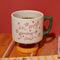 cream short ceramic mug with pink cursive 'in my growing season' with green handle and pink and yellow pedestal  on a table filled with coffee