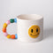 angled white ceramic mug with rainbow shapes handle and yellow smiley face on the front