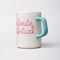 side view of ceramic mug with paper lines and pink cursive 'infinite potenial' typography across the front and light blue handle