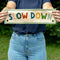 model holding beige canvas banner with colorful 'SLOW DOWN' and sage green trim