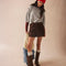model wearing tall dark brown leather boots with silver buckle and strap detail around the ankle with skirt and sweater