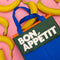 blue, green, and orange color block lunch bag with 'bon appetit' across the front on a pink ground with bananas scattered around