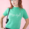 model wearing pale green short sleeve mockneck sweater with white cursive 'stay soft' across the front