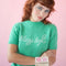 model wearing pale green short sleeve mockneck sweater with white cursive 'stay soft' across the front