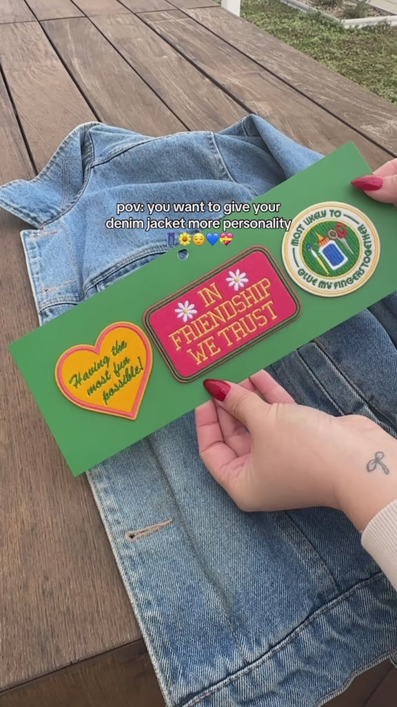 model puttingset of 3 embroidered patch stickers: yellow heart with green 'having the most fun possible' across the front, white round 'most likely to glue my fingers together' and red rectangular 'in friendship we trust' onto a denim jacket