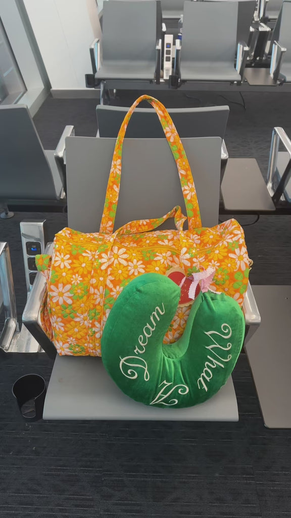 model showcasing yellow duffel bag with retro green, white and orange floral print and green travel neck pillow with white 'what a dream' across the front