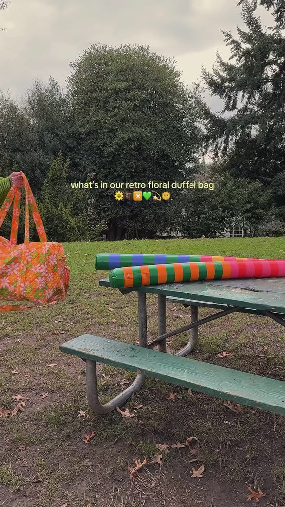 model pulling colorful items out of orange quilted duffel bag with handles and adjustable strap and has an all over yellow, white, and green retro floral print